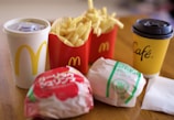 A fast food meal consisting of two burgers wrapped in branded paper, a portion of fries in a red container, a soft drink in a white cup with a yellow logo, and a yellow coffee cup branded with 'Café' on a wooden table.