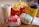 A fast food meal consisting of two burgers wrapped in branded paper, a portion of fries in a red container, a soft drink in a white cup with a yellow logo, and a yellow coffee cup branded with 'Café' on a wooden table.
