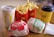 A fast food meal consisting of two burgers wrapped in branded paper, a portion of fries in a red container, a soft drink in a white cup with a yellow logo, and a yellow coffee cup branded with 'Café' on a wooden table.