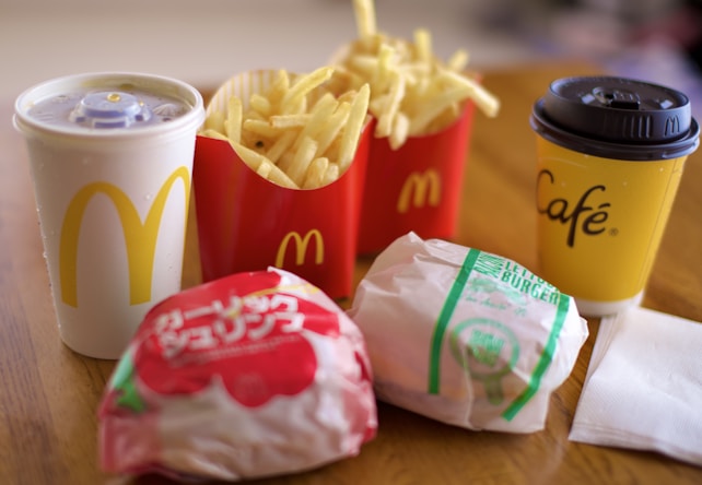 A fast food meal consisting of two burgers wrapped in branded paper, a portion of fries in a red container, a soft drink in a white cup with a yellow logo, and a yellow coffee cup branded with 'Caf&eacute;' on a wooden table.