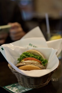 A gourmet burger is placed in a wicker basket lined with white paper. The burger is layered with fresh lettuce, a thick tomato slice, onions, and a juicy burger patty. In the background, a person holds a mobile phone next to a glass of a drink with a lemon slice.