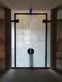 A large, modern, stone-textured door with a prominent black circular handle situated in the center. The door is framed by dark rectangular beams and bordered by two cylindrical columns on each side. The overall design is minimalist with a symmetrical layout.
