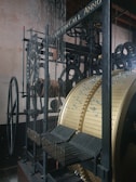 An intricate mechanical structure featuring large gears, wheels, and a cylindrical drum with perforations. The machinery appears to be part of a historical clock or automated instrument, with various metal rods and components arranged precisely. The materials include metal and wood, with a visible text inscription on one of the beams.