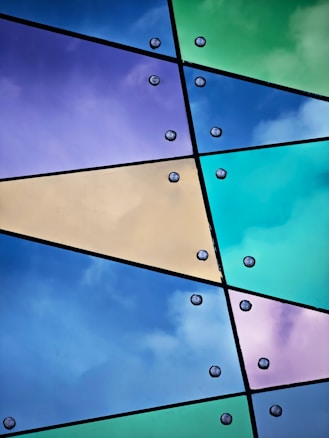 Colorful glass panels intersect with black lines, forming geometric shapes. Each panel displays vibrant shades of blue, purple, green, and yellow. Metallic rivets are visible at intersecting points, enhancing the modern and artistic appearance.