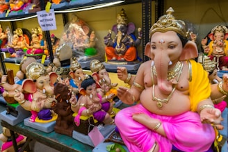 A collection of colorful Ganesha idols is displayed on shelves. The prominent idol in the foreground is adorned with a golden crown and jewelry, wearing a yellow and pink cloth. Smaller idols surround it, each uniquely decorated. A sign in the background reads 'Please do not touch the idols.' The setting appears to be a shop or market.