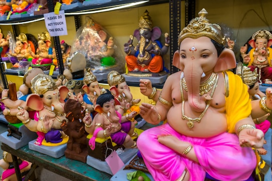 A collection of colorful Ganesha idols is displayed on shelves. The prominent idol in the foreground is adorned with a golden crown and jewelry, wearing a yellow and pink cloth. Smaller idols surround it, each uniquely decorated. A sign in the background reads 'Please do not touch the idols.' The setting appears to be a shop or market.