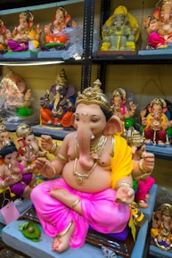 A collection of colorful, ornate statues depicting the Hindu deity Ganesha, displayed on shelves. The central figure is a large Ganesha statue, adorned with gold jewelry and a yellow and pink cloth.