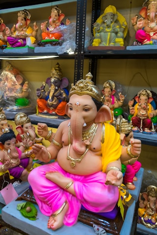 A collection of colorful, ornate statues depicting the Hindu deity Ganesha, displayed on shelves. The central figure is a large Ganesha statue, adorned with gold jewelry and a yellow and pink cloth.