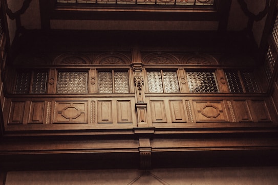 An ornate wooden balcony with intricate carvings and patterned glass panels. The woodwork features geometric shapes and decorative details, creating a sense of sophistication and historical elegance.