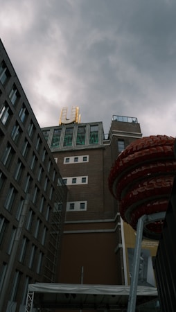 The image captures an urban scene featuring a tall brick building with a large neon U sign on top. The foreground includes a red spiral structure standing on a pole. The sky is overcast, casting a moody ambiance over the scene.