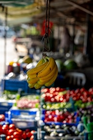 A ripe bunch of bananas hangs prominently in the foreground, with a blurred background featuring various colorful fruits and vegetables on display. The setting appears to be an outdoor market with natural light illuminating the produce.