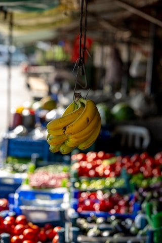 A ripe bunch of bananas hangs prominently in the foreground, with a blurred background featuring various colorful fruits and vegetables on display. The setting appears to be an outdoor market with natural light illuminating the produce.