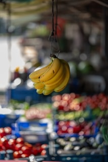 A cluster of ripe bananas hangs prominently in the foreground with a vibrant and colorful fruit market in the blurred background. Various fruits in crates can be seen, including tomatoes and possibly other produce, under soft daylight.