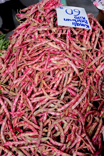 A large pile of fresh, mottled pink and green beans with a handwritten sign displaying a price. The beans are heaped in an abundant display, likely at a market setting.