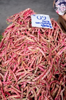 A large pile of red speckled beans is displayed, with a handwritten price sign marked '60' on top. The background is a light grey, and part of a person can be seen on the right side.