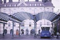A bus terminal featuring a blue bus under a modern, arched glass canopy. The background displays an ornate, historic building with several shops such as Subway. The scene includes a few pedestrians and bicycles.