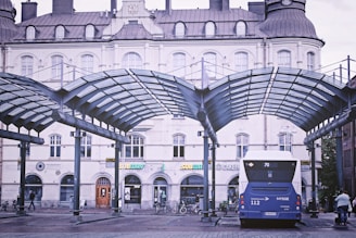 A bus terminal featuring a blue bus under a modern, arched glass canopy. The background displays an ornate, historic building with several shops such as Subway. The scene includes a few pedestrians and bicycles.
