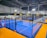 An indoor sports facility featuring several padel courts with blue playing surfaces and orange sidelines. There are players engaged in a game on one of the courts, while the others appear empty. The area is well-lit with overhead lighting and enclosed by a high ceiling and metal structure.
