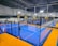An indoor sports facility featuring several padel courts with blue playing surfaces and orange sidelines. There are players engaged in a game on one of the courts, while the others appear empty. The area is well-lit with overhead lighting and enclosed by a high ceiling and metal structure.