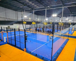 An indoor sports facility featuring several padel courts with blue playing surfaces and orange sidelines. There are players engaged in a game on one of the courts, while the others appear empty. The area is well-lit with overhead lighting and enclosed by a high ceiling and metal structure.