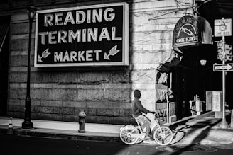 A black-and-white street scene featuring a bicyclist wearing a hoodie riding past the iconic Reading Terminal Market sign. The setting includes the corner of a building with a vintage feel, illuminated by streetlights and sunlight. Nearby, a cone and a one-way street sign contribute to the urban environment.