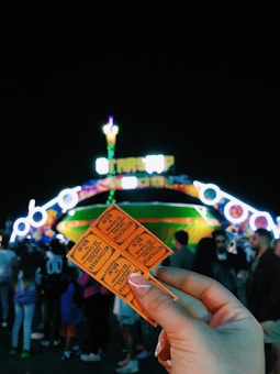 Several orange tickets are held in a hand in the foreground while a lively, brightly lit amusement park ride is in the background. The surrounding area is filled with a crowd of people, and the night atmosphere is darkly contrasted with colorful lights.