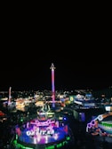 A brightly lit amusement park at night, featuring numerous colorful rides and attractions. The centerpiece is a tall, illuminated ride that stands out against the dark sky. Crowds of people are scattered throughout the park, enjoying the vibrant atmosphere.