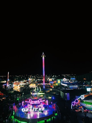 A brightly lit amusement park at night, featuring numerous colorful rides and attractions. The centerpiece is a tall, illuminated ride that stands out against the dark sky. Crowds of people are scattered throughout the park, enjoying the vibrant atmosphere.