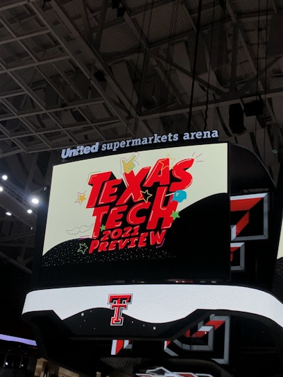 A scoreboard displaying the text 'Texas Tech 2021 Preview' in bold red letters with decorative elements like stars and clouds. The background is predominantly dark with bright accent colors, and the setting is within an indoor sports arena, specifically the United Supermarkets Arena.