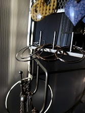 A metal jewelry stand holds several pairs of earrings in various shapes and materials. The earrings include circular metal hoops and a pair of yellow checkered and blue marbled designs. The stand is positioned against a textured wall, with soft lighting creating shadows and highlighting the metallic surfaces.