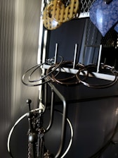 A metal jewelry stand holds several pairs of earrings in various shapes and materials. The earrings include circular metal hoops and a pair of yellow checkered and blue marbled designs. The stand is positioned against a textured wall, with soft lighting creating shadows and highlighting the metallic surfaces.