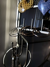 A metal jewelry stand holds several pairs of earrings in various shapes and materials. The earrings include circular metal hoops and a pair of yellow checkered and blue marbled designs. The stand is positioned against a textured wall, with soft lighting creating shadows and highlighting the metallic surfaces.
