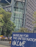 A modern architectural building displaying the BBVA logo with a glass facade, surrounded by lush green trees. In the foreground, a blue container with the words 'Museum for the United Nations UN.Live' is visible, and there are people walking on a pathway nearby.
