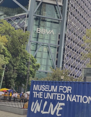 A modern architectural building displaying the BBVA logo with a glass facade, surrounded by lush green trees. In the foreground, a blue container with the words 'Museum for the United Nations UN.Live' is visible, and there are people walking on a pathway nearby.