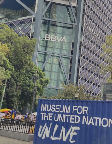 A modern architectural building displaying the BBVA logo with a glass facade, surrounded by lush green trees. In the foreground, a blue container with the words 'Museum for the United Nations UN.Live' is visible, and there are people walking on a pathway nearby.