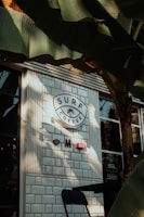 A café exterior with white brick tiles and a round sign that reads 'Surf Coffee' with a palm tree icon. There are a few decorative signs on the wall under the main sign. Large green banana leaves partially cover the view, casting shadows on the café wall and window. A black chair can be seen in the lower part of the image.