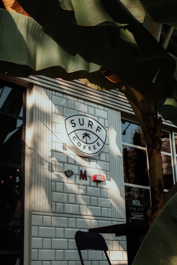 A café exterior with white brick tiles and a round sign that reads 'Surf Coffee' with a palm tree icon. There are a few decorative signs on the wall under the main sign. Large green banana leaves partially cover the view, casting shadows on the café wall and window. A black chair can be seen in the lower part of the image.