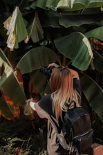 A person with long hair and a black backpack is taking a photograph using a camera, surrounded by large, lush green tropical leaves. Sunlight is filtering through the leaves, creating patches of light and shadow.
