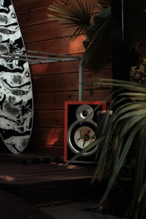 A surfboard with a black and white marbled design leans against a wooden wall. In front of it, a large speaker with a red frame sits on a wooden deck. The area is surrounded by lush green palm leaves, casting shadows on the wooden panels.
