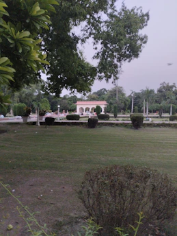 A well-maintained garden with lush green grass and neatly trimmed bushes. In the background, there is a light-colored decorative structure with arches, surrounded by more greenery. The sky appears overcast and there are tall trees with dense foliage.