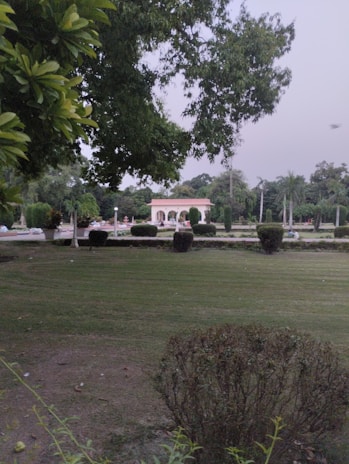 A well-maintained garden with lush green grass and neatly trimmed bushes. In the background, there is a light-colored decorative structure with arches, surrounded by more greenery. The sky appears overcast and there are tall trees with dense foliage.