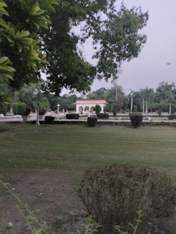 A well-maintained garden with lush green grass and neatly trimmed bushes. In the background, there is a light-colored decorative structure with arches, surrounded by more greenery. The sky appears overcast and there are tall trees with dense foliage.