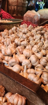 A wooden crate filled with numerous garlic bulbs is situated in a market setting. In the background, there are baskets containing red onions and other produce. The setting suggests a bustling, vibrant market environment.