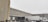 Snow falls gently in an industrial loading dock area with two large trucks parked against a warehouse building. The ground and the trucks are partially covered with snow, and the sky is overcast, creating a cold and wintry atmosphere.