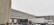 Snow falls gently in an industrial loading dock area with two large trucks parked against a warehouse building. The ground and the trucks are partially covered with snow, and the sky is overcast, creating a cold and wintry atmosphere.