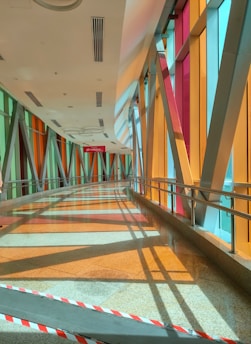 A brightly lit hallway features large, colorful glass panels on one side, casting vivid reflections and shadows on the floor. The ceiling has sleek, modern lighting, and metal railings run along the walls. A red and white striped safety barrier is positioned at the entrance.