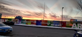 A colorful wall with promotional text and graphics lines a street at sunset. The sky is filled with dramatic clouds reflecting the colors of the setting sun. There are several parked cars and streetlights, and a building with illuminated windows is visible in the background.