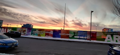 A colorful wall with promotional text and graphics lines a street at sunset. The sky is filled with dramatic clouds reflecting the colors of the setting sun. There are several parked cars and streetlights, and a building with illuminated windows is visible in the background.