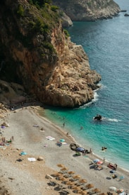 A beautiful rocky coastline with turquoise waters where people are enjoying sunbathing and swimming. The beach is lined with pebbles, and there are sun umbrellas and beach towels scattered along the shore. The landscape is dominated by steep cliffs with lush greenery on top, adding to the natural beauty of the setting.