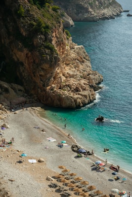 A beautiful rocky coastline with turquoise waters where people are enjoying sunbathing and swimming. The beach is lined with pebbles, and there are sun umbrellas and beach towels scattered along the shore. The landscape is dominated by steep cliffs with lush greenery on top, adding to the natural beauty of the setting.